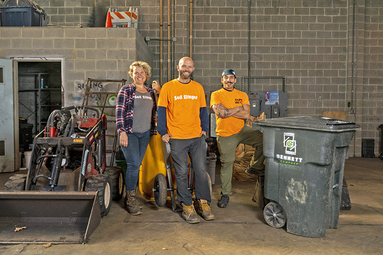 three people stand in a workshop next to trash/compost bin and small tractor