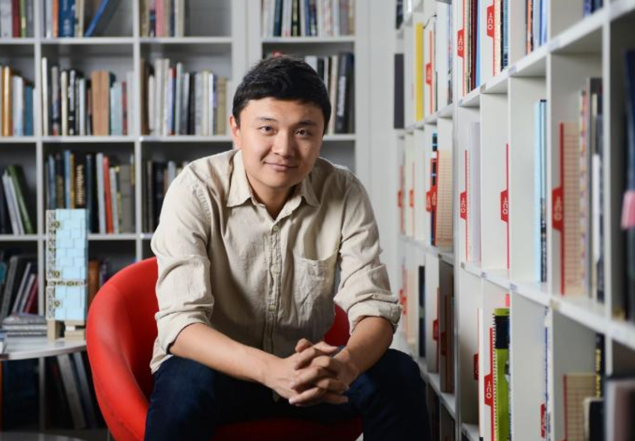 man sitting in chair, surrounded by books on shelves. 