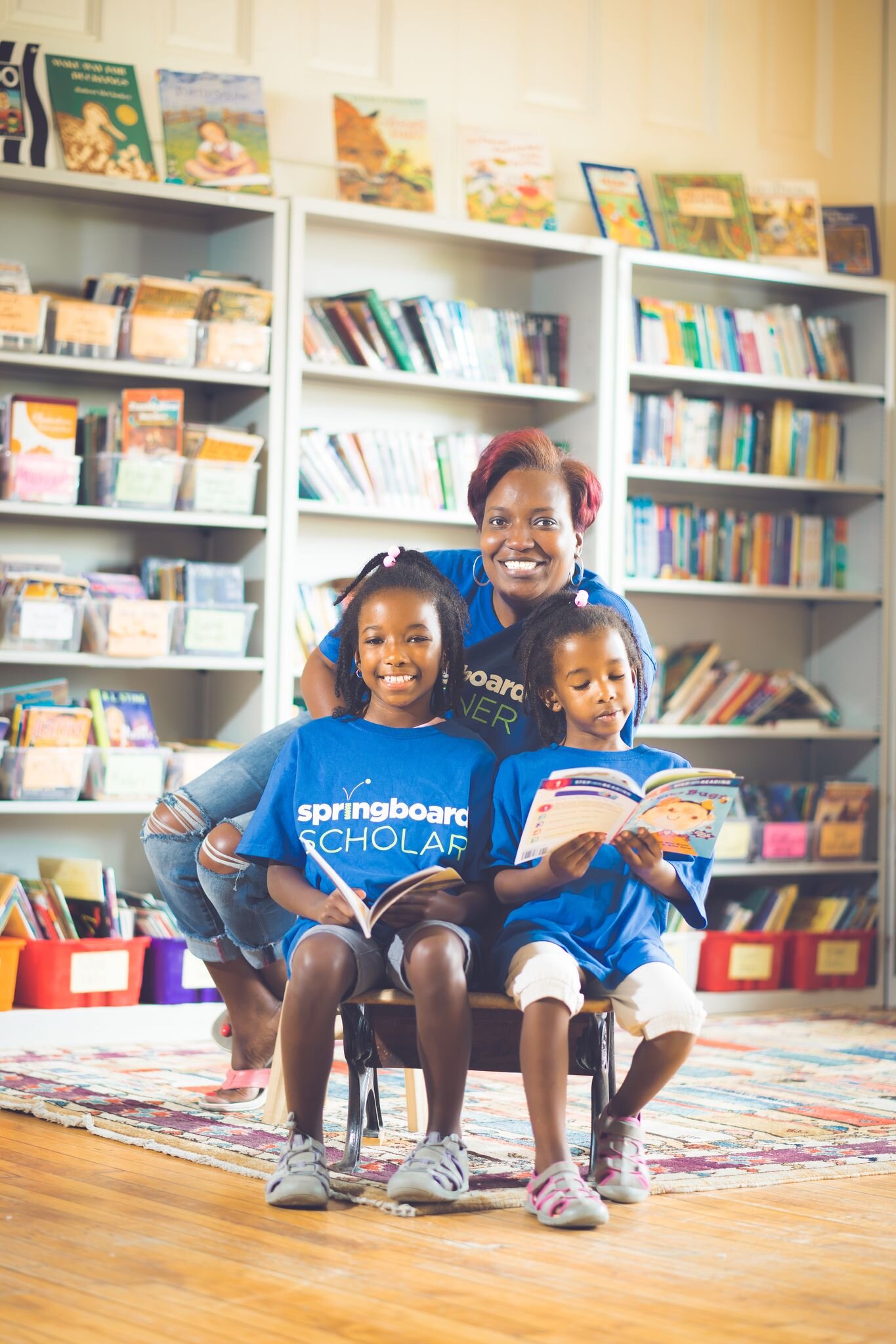 woman posing behind two children reading books with springboard scholars tee shirts, in a library
