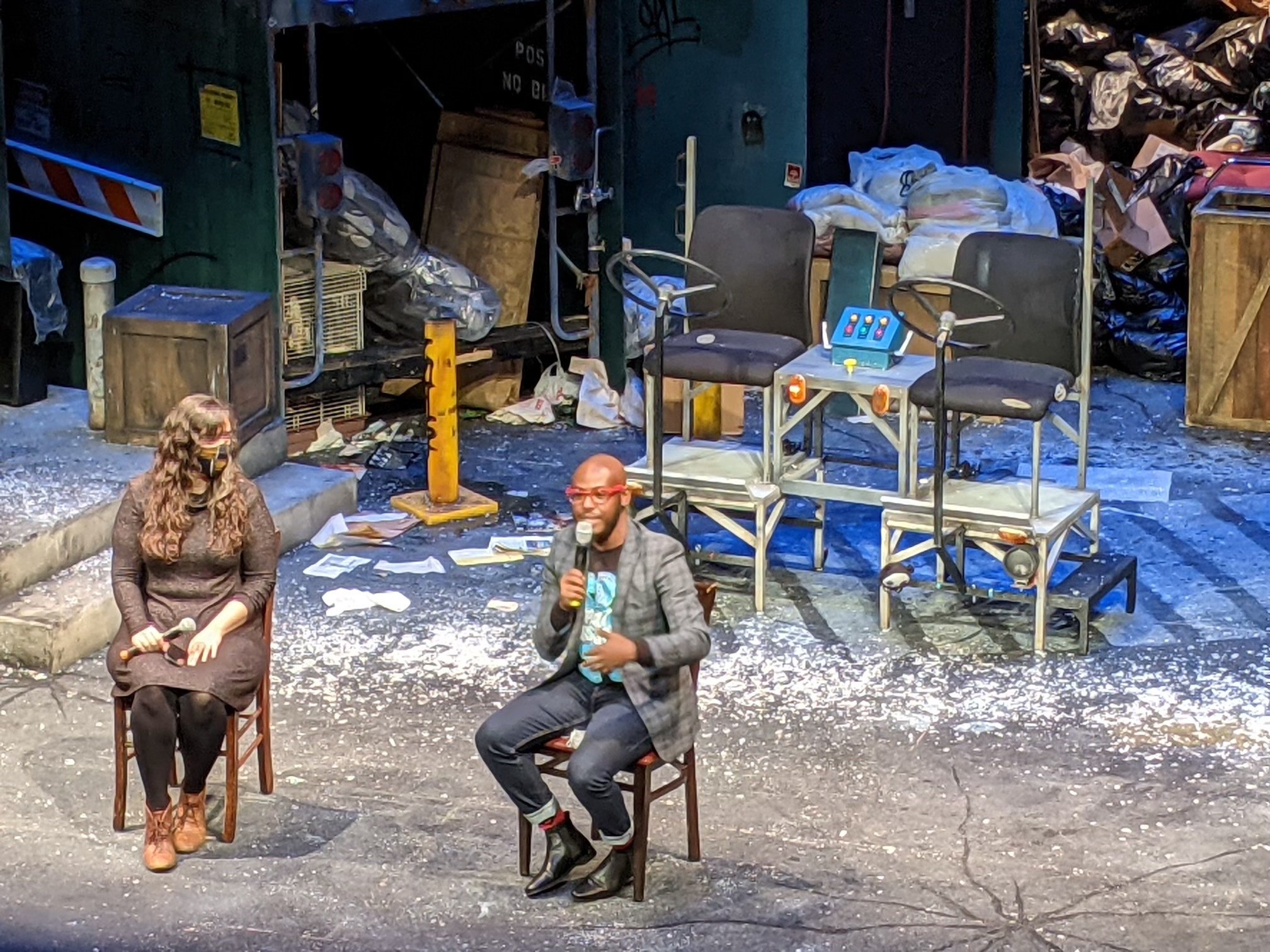 two people sit on chairs on a theater stage, one with a microphone